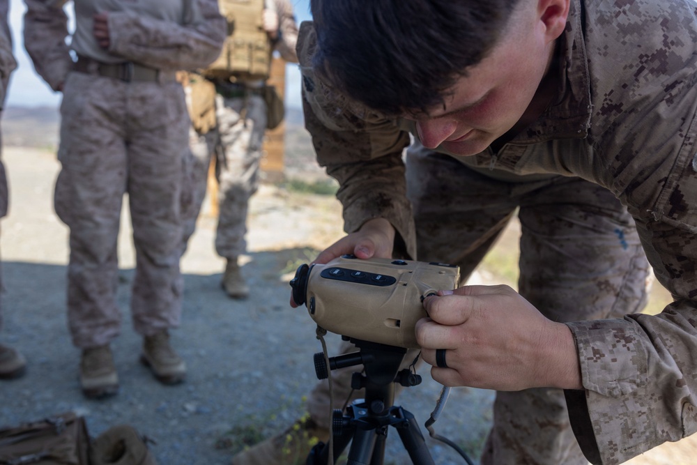 A U.S. Marine in MARPAT digital camouflage uniform is closely examining or adjusting a tan-colored military optical/laser rangefinder device mounted on a tripod. Several other Marines in similar uniforms are visible in the background observing. The setting appears to be an outdoor training or field exercise environment on rocky/gravel terrain.