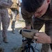 A U.S. Marine in MARPAT digital camouflage uniform is closely examining or adjusting a tan-colored military optical/laser rangefinder device mounted on a tripod. Several other Marines in similar uniforms are visible in the background observing. The setting appears to be an outdoor training or field exercise environment on rocky/gravel terrain.