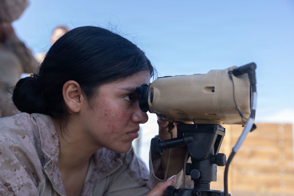 A female military service member in desert camouflage (MARPAT) uniform is looking through a tan-colored optical device mounted on a tripod. The image appears to be taken outdoors in a dry, arid environment under a clear blue sky. The subject is focused and engaged with the optical equipment, suggesting a training or operational observation activity.