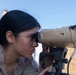 A female military service member in desert camouflage (MARPAT) uniform is looking through a tan-colored optical device mounted on a tripod. The image appears to be taken outdoors in a dry, arid environment under a clear blue sky. The subject is focused and engaged with the optical equipment, suggesting a training or operational observation activity.