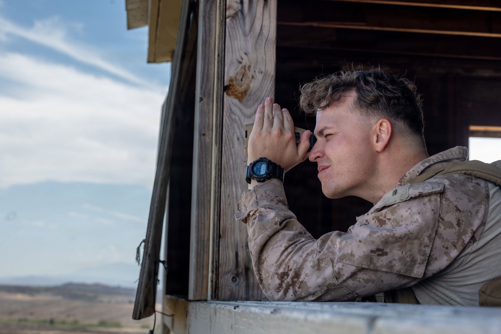 A military service member in desert camouflage (MARPAT) uniform is using a compact optical device, likely a rangefinder or monocular, while leaning out of a wooden observation post window. The arid, open terrain visible in the background suggests a training environment or forward operating position. The image captures a surveillance or reconnaissance activity in a desert setting.