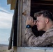 A military service member in desert camouflage (MARPAT) uniform is using a compact optical device, likely a rangefinder or monocular, while leaning out of a wooden observation post window. The arid, open terrain visible in the background suggests a training environment or forward operating position. The image captures a surveillance or reconnaissance activity in a desert setting.