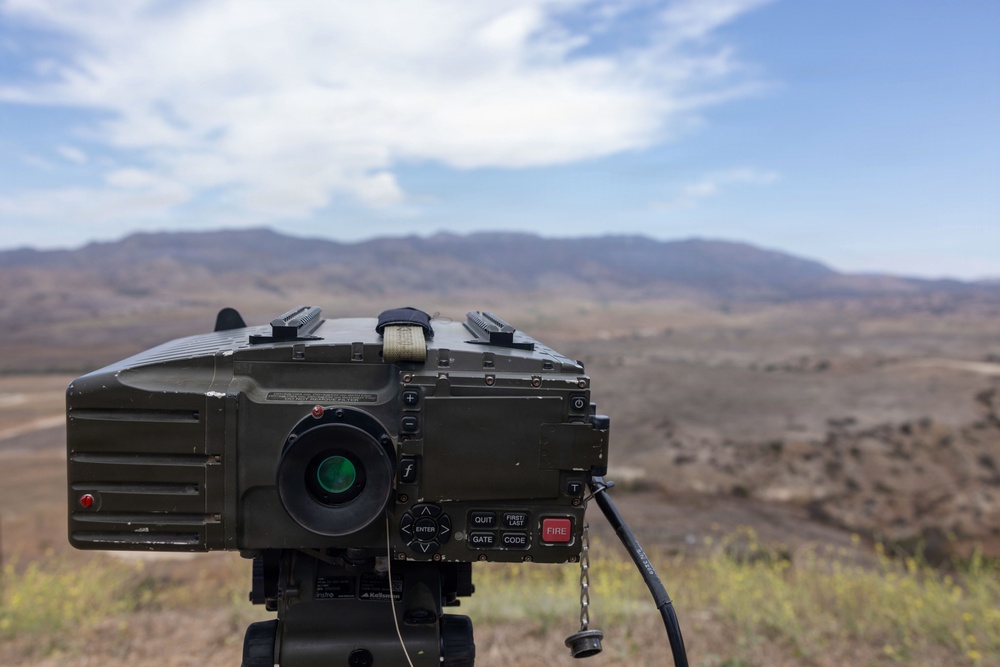 A military optical/targeting device, likely a laser rangefinder or designator system, is mounted on a tripod in an arid, desert-like terrain with mountains visible in the background. The device is olive drab in color and features multiple control buttons including a prominently labeled red 'FIRE' button, along with labels such as 'QUIT', 'GATE', 'FIRST/LAST', and 'CODE'. The background shows a semi-arid landscape under a partly cloudy sky.