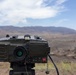 A military optical/targeting device, likely a laser rangefinder or designator system, is mounted on a tripod in an arid, desert-like terrain with mountains visible in the background. The device is olive drab in color and features multiple control buttons including a prominently labeled red 'FIRE' button, along with labels such as 'QUIT', 'GATE', 'FIRST/LAST', and 'CODE'. The background shows a semi-arid landscape under a partly cloudy sky.