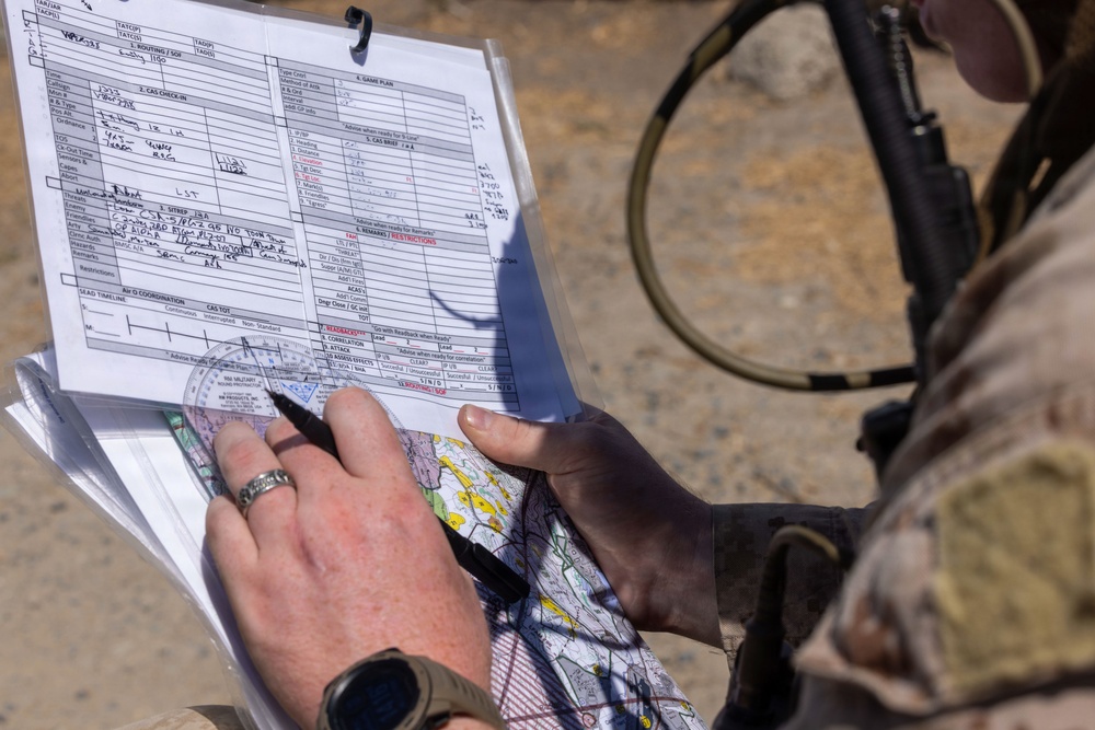 A military personnel in camouflage uniform is using a protractor/plotter tool on an aeronautical or topographic map while referencing a filled-out military tactical form, likely a CAS (Close Air Support) 9-line brief card. The individual is wearing a watch and a ring, and has a weapon slung over their shoulder. The setting appears to be an outdoor field environment with sandy/arid terrain visible in the background.