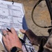A military personnel in camouflage uniform is using a protractor/plotter tool on an aeronautical or topographic map while referencing a filled-out military tactical form, likely a CAS (Close Air Support) 9-line brief card. The individual is wearing a watch and a ring, and has a weapon slung over their shoulder. The setting appears to be an outdoor field environment with sandy/arid terrain visible in the background.