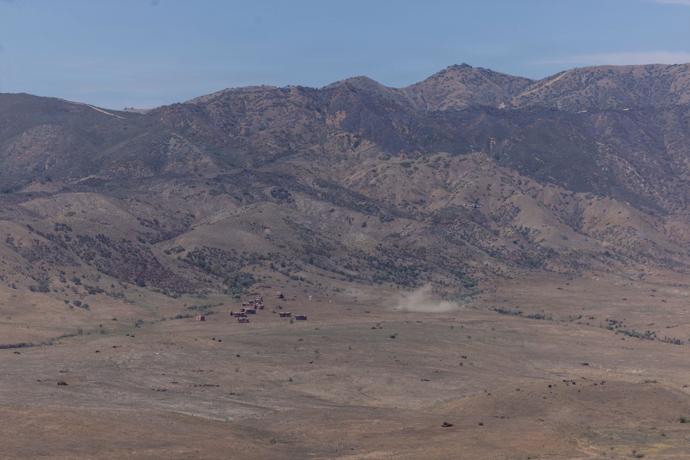 A wide-angle landscape photograph showing an arid, semi-desert valley with a cluster of small reddish-brown structures in the middle distance. A dust cloud is visible near the structures, and what appears to be a helicopter or small aircraft is flying nearby. A rugged mountain range forms the backdrop under a partly cloudy blue sky.