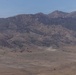 A wide-angle landscape photograph showing an arid, semi-desert valley with a cluster of small reddish-brown structures in the middle distance. A dust cloud is visible near the structures, and what appears to be a helicopter or small aircraft is flying nearby. A rugged mountain range forms the backdrop under a partly cloudy blue sky.