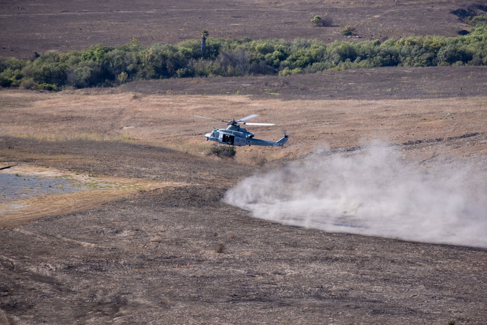 A military helicopter, appearing to be a UH-1Y Venom or similar utility helicopter in light gray/blue livery, is flying at low altitude over an arid, open field. The rotor wash is kicking up a large dust cloud from the dry ground below. The surrounding terrain includes dry grassland and scrub vegetation in the background.