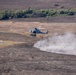 A military helicopter, appearing to be a UH-1Y Venom or similar utility helicopter in light gray/blue livery, is flying at low altitude over an arid, open field. The rotor wash is kicking up a large dust cloud from the dry ground below. The surrounding terrain includes dry grassland and scrub vegetation in the background.