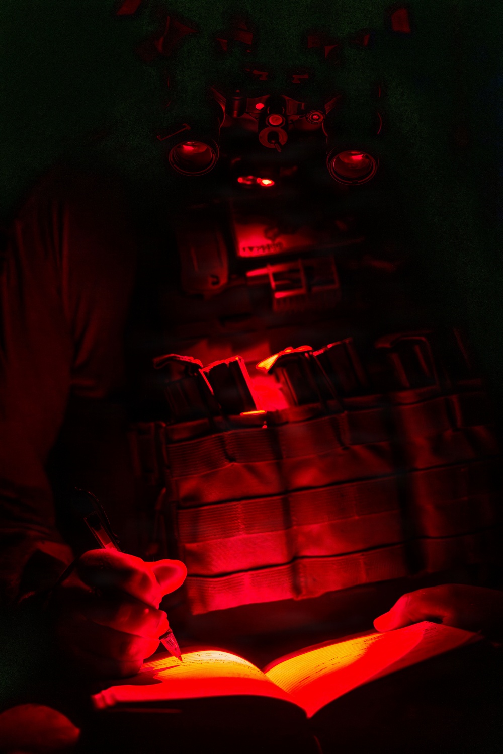 A person in full tactical/military gear is writing in a notebook or logbook under red light conditions. The scene is extremely dark with red illumination as the only light source, consistent with night operations where red light is used to preserve night vision. The individual appears to be wearing a plate carrier vest with magazine pouches and a helmet-mounted device, likely night vision goggles.