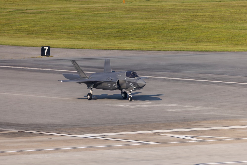 A single stealth fighter jet, identifiable as an F-35 Lightning II, is taxiing on an airport or military airfield tarmac. The aircraft is painted in a dark gray low-visibility scheme and appears to be moving along a taxiway marked with the number '7'. The image is taken during daylight with warm, low-angle lighting suggesting early morning or late afternoon conditions.