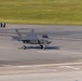 A single stealth fighter jet, identifiable as an F-35 Lightning II, is taxiing on an airport or military airfield tarmac. The aircraft is painted in a dark gray low-visibility scheme and appears to be moving along a taxiway marked with the number '7'. The image is taken during daylight with warm, low-angle lighting suggesting early morning or late afternoon conditions.