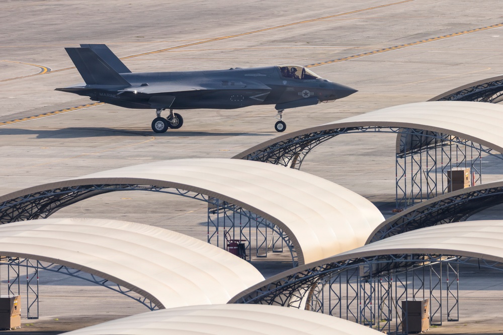A dark gray stealth fighter jet, identifiable as an F-35, is taxiing on a military airfield tarmac. The aircraft bears visible 'MARINES' markings and a star-and-bar insignia on its fuselage. In the foreground, multiple white arch-shaped aircraft shelters or soft-sided hangars are arranged in rows on the flight line.
