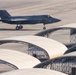 A dark gray stealth fighter jet, identifiable as an F-35, is taxiing on a military airfield tarmac. The aircraft bears visible 'MARINES' markings and a star-and-bar insignia on its fuselage. In the foreground, multiple white arch-shaped aircraft shelters or soft-sided hangars are arranged in rows on the flight line.