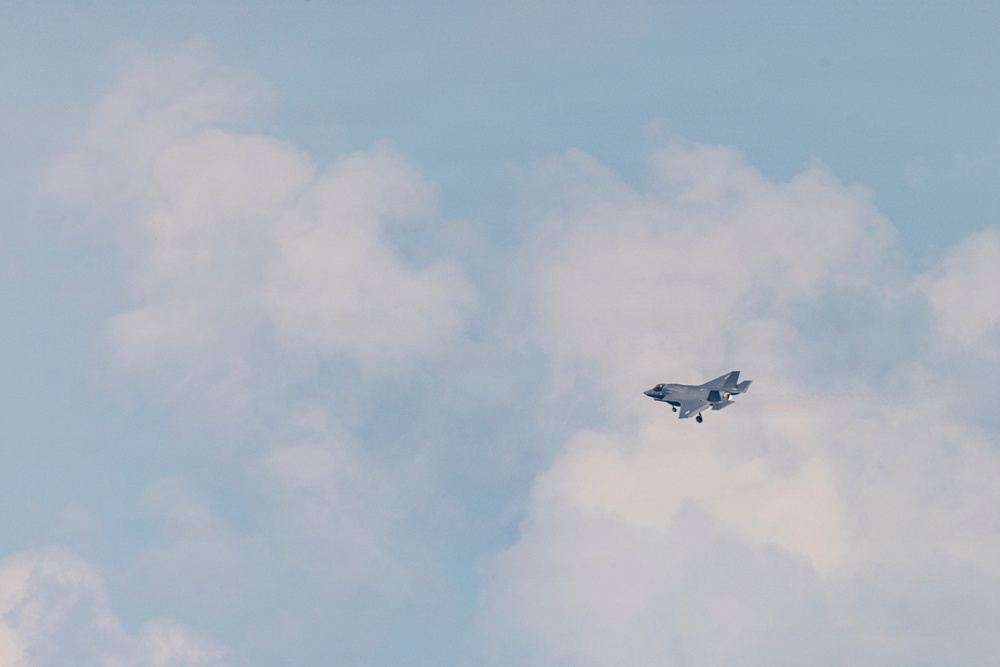 A single military jet aircraft is captured in flight against a partly cloudy sky. The aircraft appears to be a fifth-generation stealth fighter, consistent with an F-35 Lightning II, photographed at a distance. The image shows the aircraft in level flight with landing gear retracted.
