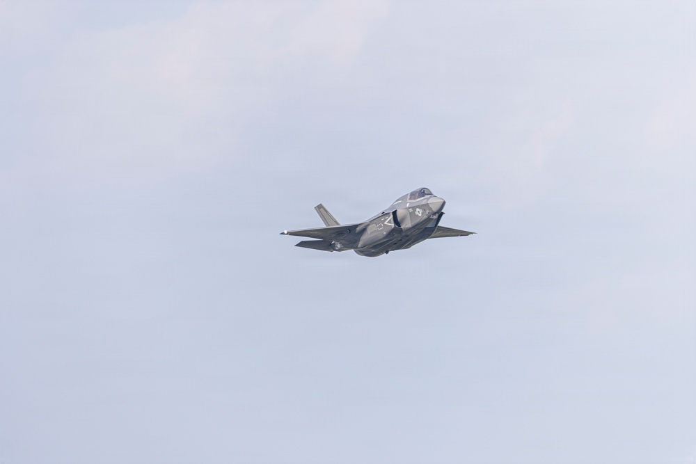 A single military stealth fighter jet is captured in flight against an overcast, pale blue-gray sky. The aircraft displays a low-observable design consistent with fifth-generation fighter characteristics, including blended surfaces and a single engine exhaust. The image appears to have been taken at an airshow or during a training flight.