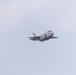 A single military stealth fighter jet is captured in flight against an overcast, pale blue-gray sky. The aircraft displays a low-observable design consistent with fifth-generation fighter characteristics, including blended surfaces and a single engine exhaust. The image appears to have been taken at an airshow or during a training flight.
