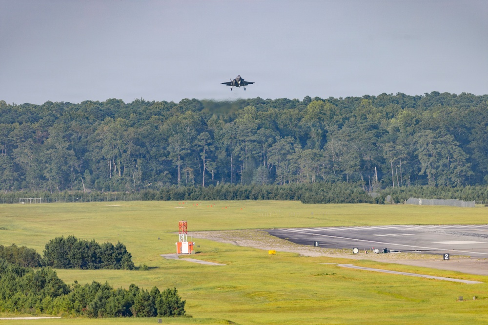 A military jet aircraft, consistent with an F-35 design, is captured in low-altitude flight approaching or departing a runway at a military airfield. The aircraft appears to be producing exhaust or heat distortion visible behind it. The airfield is surrounded by dense green tree lines with open grass infields and runway infrastructure visible in the foreground.