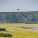 A military jet aircraft, consistent with an F-35 design, is captured in low-altitude flight approaching or departing a runway at a military airfield. The aircraft appears to be producing exhaust or heat distortion visible behind it. The airfield is surrounded by dense green tree lines with open grass infields and runway infrastructure visible in the foreground.