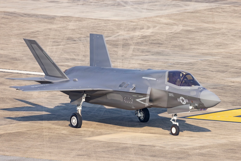 A gray stealth fighter jet is taxiing on a concrete airfield runway. The aircraft displays U.S. military markings including a star-and-bar insignia on the fuselage. A pilot is visible in the cockpit, and the aircraft appears to be in operational condition.