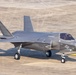 A gray stealth fighter jet is taxiing on a concrete airfield runway. The aircraft displays U.S. military markings including a star-and-bar insignia on the fuselage. A pilot is visible in the cockpit, and the aircraft appears to be in operational condition.