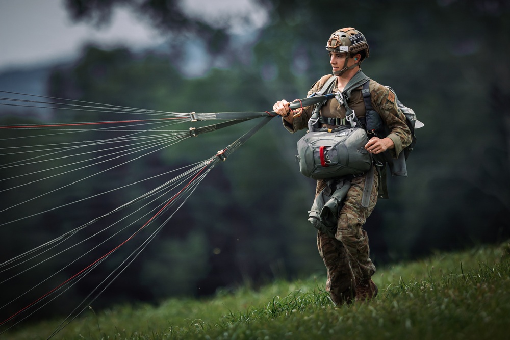 A military paratrooper is shown on a grassy field after a landing, gathering and controlling parachute suspension lines. The soldier is wearing full combat gear including a tactical helmet, multicam camouflage uniform, and a parachute harness system. The image captures the post-landing recovery phase of a parachute operation.
