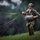 A military paratrooper is shown on a grassy field after a landing, gathering and controlling parachute suspension lines. The soldier is wearing full combat gear including a tactical helmet, multicam camouflage uniform, and a parachute harness system. The image captures the post-landing recovery phase of a parachute operation.