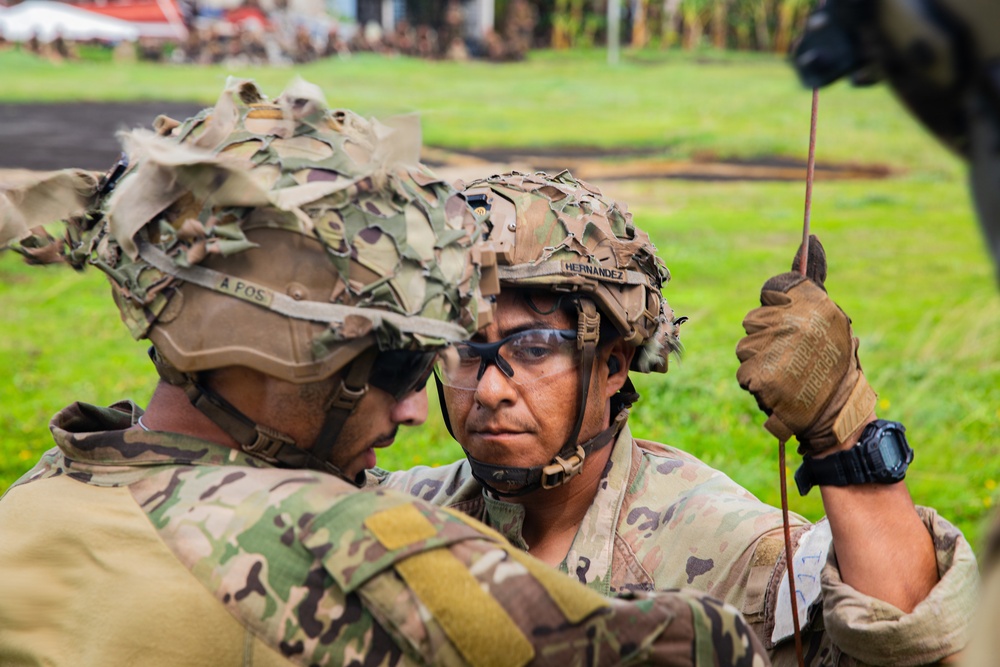 Two soldiers in full combat gear with camouflage-covered helmets are closely examining or coordinating over an object, likely during a military training exercise. The soldier facing the camera has the name tape 'HERNANDEZ' visible on their helmet and is wearing protective eyewear and tactical gloves. The setting appears to be an outdoor training area with grass and a paved surface visible in the background.