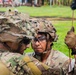 Two soldiers in full combat gear with camouflage-covered helmets are closely examining or coordinating over an object, likely during a military training exercise. The soldier facing the camera has the name tape 'HERNANDEZ' visible on their helmet and is wearing protective eyewear and tactical gloves. The setting appears to be an outdoor training area with grass and a paved surface visible in the background.