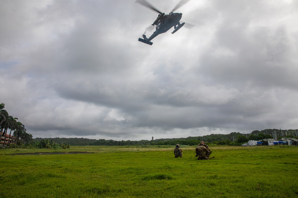 Two military personnel in camouflage uniforms are kneeling on a green grassy field while a military helicopter hovers overhead at low altitude. The scene appears to depict a tactical training exercise or insertion/extraction operation in a tropical environment. Buildings and palm trees are visible in the background under an overcast sky.