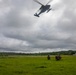Two military personnel in camouflage uniforms are kneeling on a green grassy field while a military helicopter hovers overhead at low altitude. The scene appears to depict a tactical training exercise or insertion/extraction operation in a tropical environment. Buildings and palm trees are visible in the background under an overcast sky.