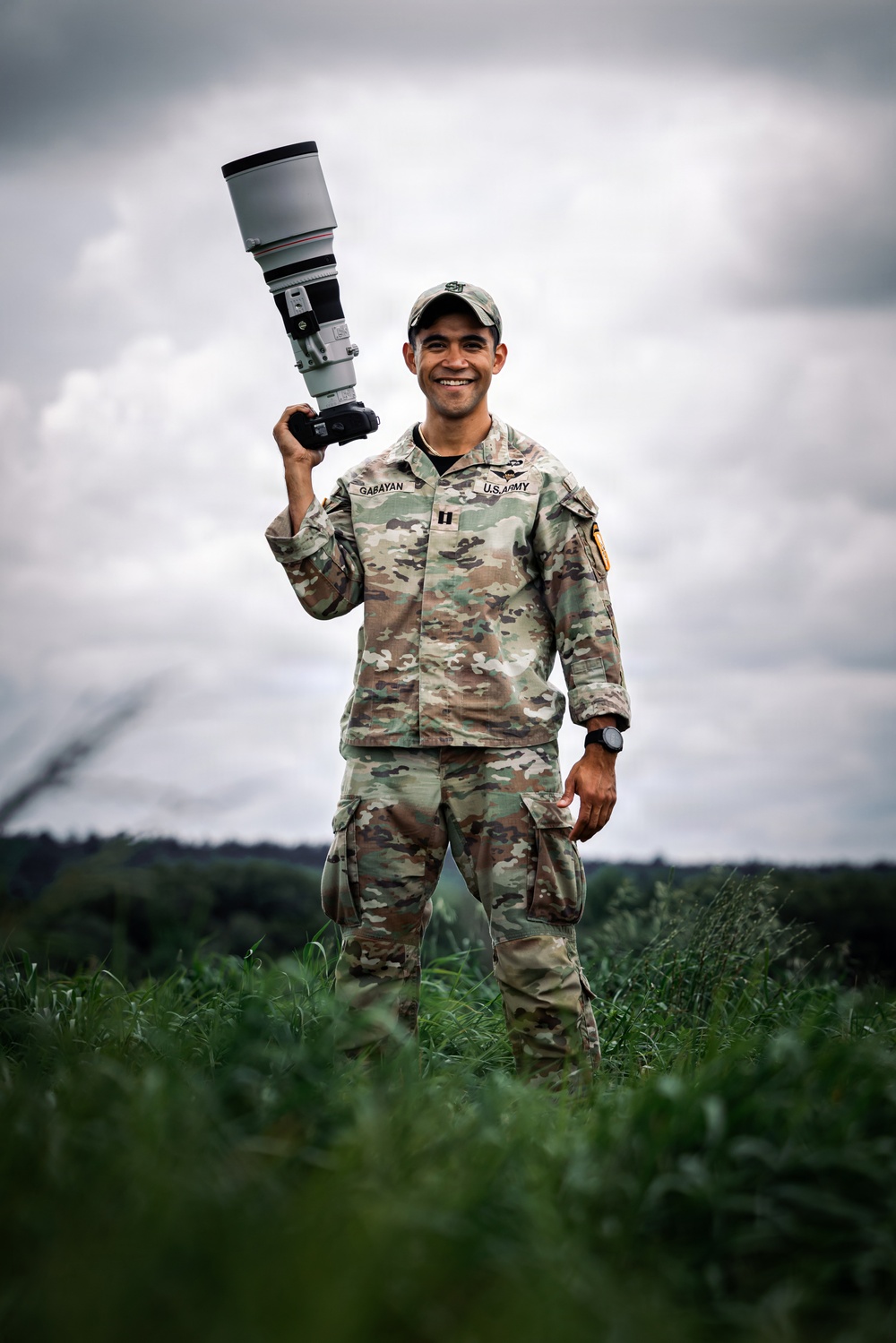 A U.S. Army soldier named Gabayan, holding a large telephoto camera lens/DSLR camera raised above shoulder height, stands smiling in an outdoor field setting. He is wearing OCP (Operational Camouflage Pattern) uniform with a captain's rank insignia and a baseball-style patrol cap. The image appears to be a professional portrait taken in a green, grassy area under an overcast sky.
