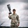 A U.S. Army soldier named Gabayan, holding a large telephoto camera lens/DSLR camera raised above shoulder height, stands smiling in an outdoor field setting. He is wearing OCP (Operational Camouflage Pattern) uniform with a captain's rank insignia and a baseball-style patrol cap. The image appears to be a professional portrait taken in a green, grassy area under an overcast sky.