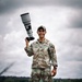 A U.S. Army soldier named Gabayan, holding a large telephoto camera lens/DSLR camera raised above shoulder height, stands smiling in an outdoor field setting. He is wearing OCP (Operational Camouflage Pattern) uniform with a captain's rank insignia and a baseball-style patrol cap. The image appears to be a professional portrait taken in a green, grassy area under an overcast sky.