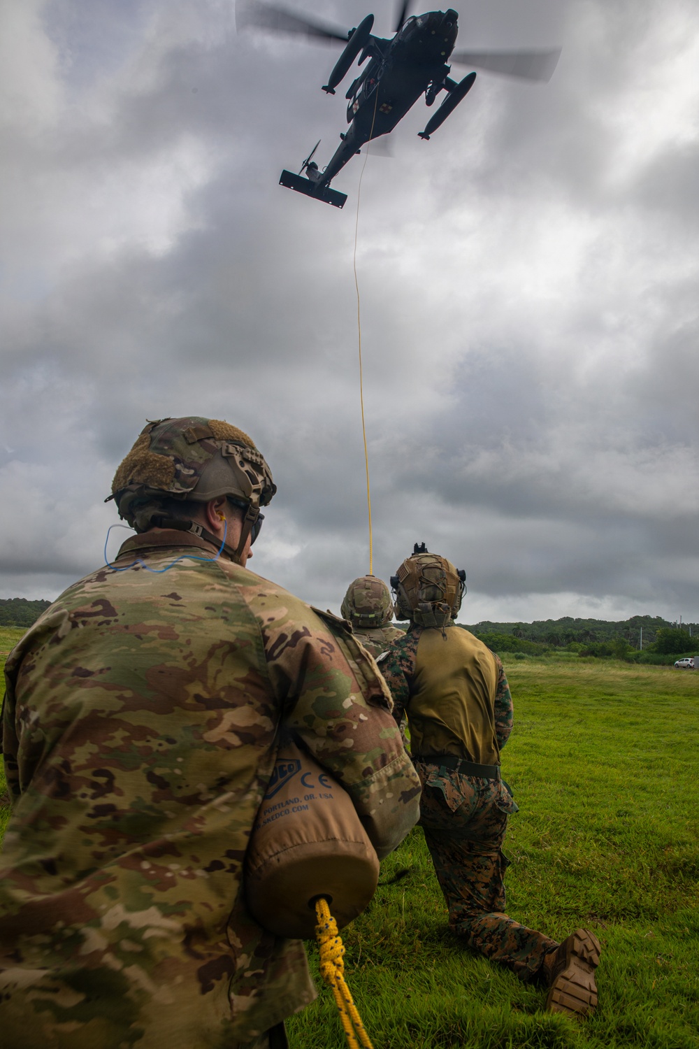Military personnel on a grassy field are engaged in a helicopter hoist or extraction exercise, holding a yellow rope connected to a hovering helicopter above. At least three service members in different camouflage uniforms are visible, suggesting a joint or combined arms training operation. The overcast sky and lush green surroundings suggest a tropical or subtropical environment.