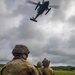 Military personnel on a grassy field are engaged in a helicopter hoist or extraction exercise, holding a yellow rope connected to a hovering helicopter above. At least three service members in different camouflage uniforms are visible, suggesting a joint or combined arms training operation. The overcast sky and lush green surroundings suggest a tropical or subtropical environment.