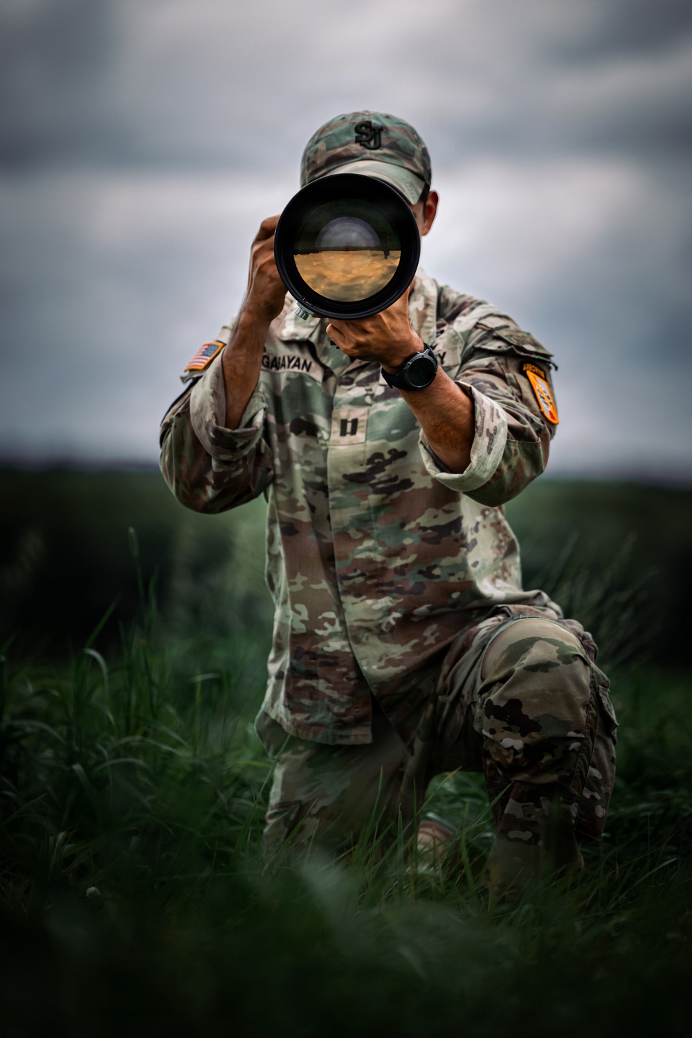 A U.S. Army soldier in OCP (Operational Camouflage Pattern) uniform is kneeling in a grassy field while holding a large telephoto camera lens up to their face. The soldier's name tape reads 'GAIAYAN' and rank insignia indicates a Captain (two vertical bars). The image is a high-quality, dramatic photograph taken under overcast skies.