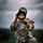 A U.S. Army soldier in OCP (Operational Camouflage Pattern) uniform is kneeling in a grassy field while holding a large telephoto camera lens up to their face. The soldier's name tape reads 'GAIAYAN' and rank insignia indicates a Captain (two vertical bars). The image is a high-quality, dramatic photograph taken under overcast skies.