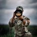 A U.S. Army soldier in OCP (Operational Camouflage Pattern) uniform is kneeling in a grassy field while holding a large telephoto camera lens up to their face. The soldier's name tape reads 'GAIAYAN' and rank insignia indicates a Captain (two vertical bars). The image is a high-quality, dramatic photograph taken under overcast skies.