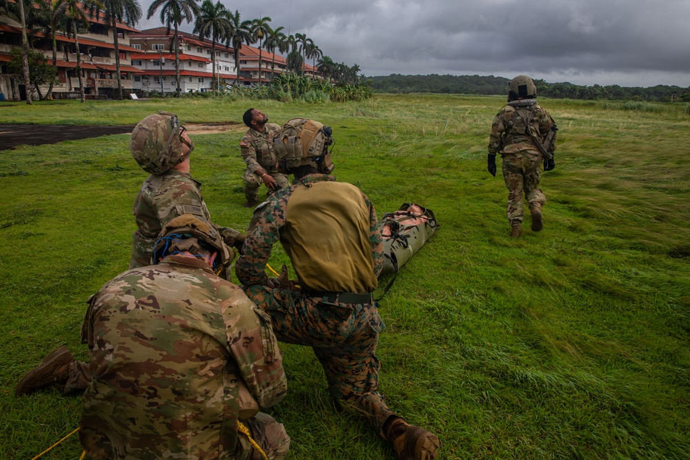 Military personnel in camouflage uniforms are conducting a casualty evacuation (CASEVAC) training exercise on a grassy field. Multiple soldiers are working together to carry a simulated casualty on a litter/stretcher, while one soldier walks ahead. The setting appears to be a tropical location, evidenced by palm trees and a multi-story building in the background.