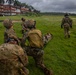 Military personnel in camouflage uniforms are conducting a casualty evacuation (CASEVAC) training exercise on a grassy field. Multiple soldiers are working together to carry a simulated casualty on a litter/stretcher, while one soldier walks ahead. The setting appears to be a tropical location, evidenced by palm trees and a multi-story building in the background.