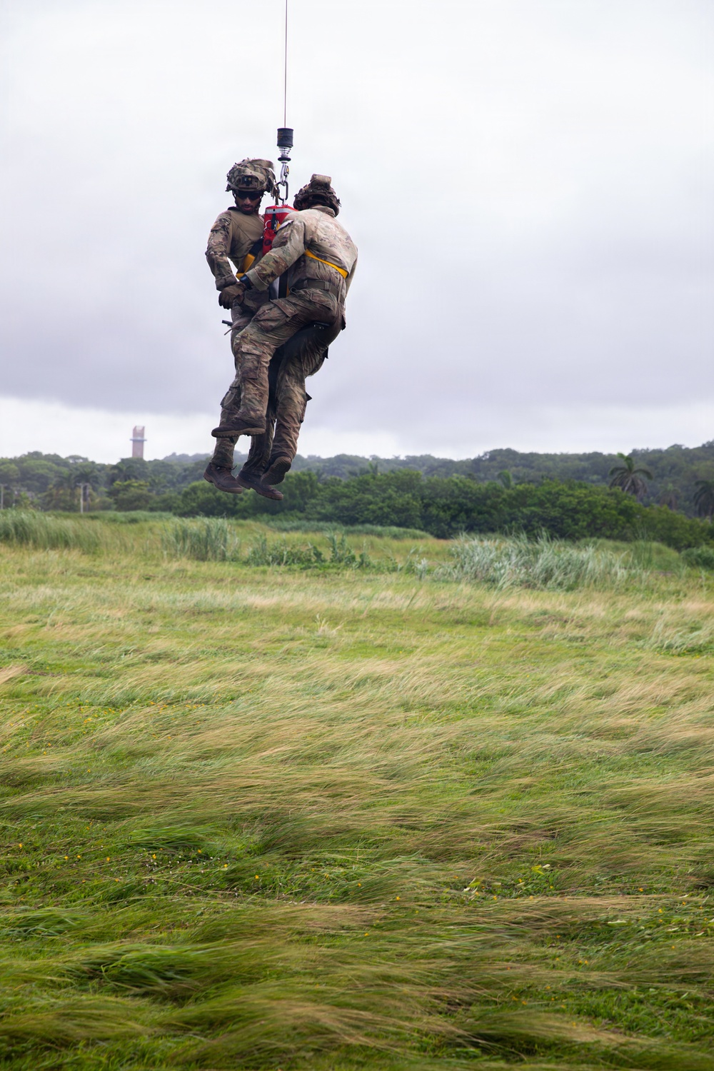 Two military personnel in camouflage uniforms are suspended mid-air via a hoist cable, likely from a helicopter above the frame. One individual appears to be a rescue operator holding or securing a second person, who is wearing a red rescue harness with yellow markings. The scene takes place over a wind-swept grassy field with dense tree line and a structure visible in the background.