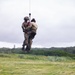 Two military personnel in camouflage uniforms are suspended mid-air via a hoist cable, likely from a helicopter above the frame. One individual appears to be a rescue operator holding or securing a second person, who is wearing a red rescue harness with yellow markings. The scene takes place over a wind-swept grassy field with dense tree line and a structure visible in the background.