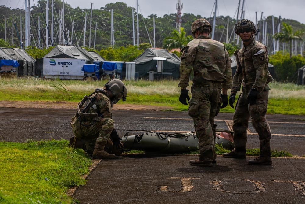 Three military personnel in camouflage uniforms and helmets are working with what appears to be a military litter or stretcher on a paved surface. The setting appears to be a forward operating base or staging area with military tents, equipment containers, and a marina with sailboat masts visible in the background. The environment suggests a tropical or subtropical location based on the vegetation.