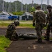 Three military personnel in camouflage uniforms and helmets are working with what appears to be a military litter or stretcher on a paved surface. The setting appears to be a forward operating base or staging area with military tents, equipment containers, and a marina with sailboat masts visible in the background. The environment suggests a tropical or subtropical location based on the vegetation.