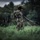 A military paratrooper in full camouflage gear stands in a grassy field after an apparent landing, gathering parachute lines. The soldier is wearing a tactical helmet, harness, and digital camouflage uniform, and appears to be a female service member based on visible facial features. The overcast sky and lush green treeline in the background suggest a training or operational environment.
