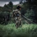 A military paratrooper in full camouflage gear stands in a grassy field after an apparent landing, gathering parachute lines. The soldier is wearing a tactical helmet, harness, and digital camouflage uniform, and appears to be a female service member based on visible facial features. The overcast sky and lush green treeline in the background suggest a training or operational environment.