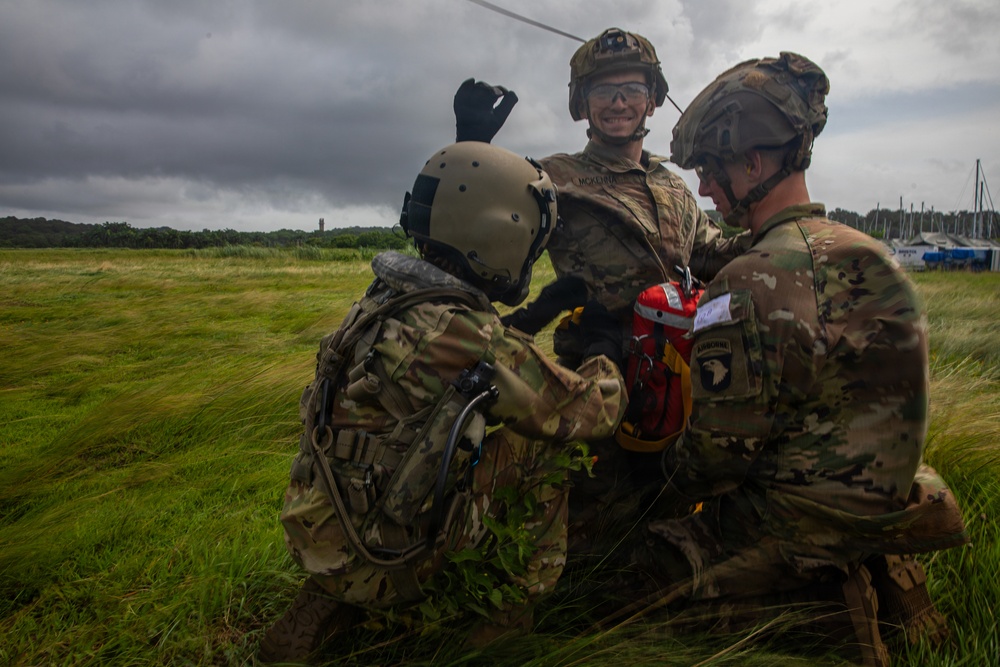 Three military personnel in MultiCam/OCP camouflage uniforms are gathered on a grassy field under overcast skies, appearing to handle equipment including a red bag or container. One soldier is smiling and wearing clear protective eyewear and a combat helmet, while another wears an aviation-style helmet. A 101st Airborne Division patch is visible on one soldier's right shoulder.