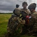Three military personnel in MultiCam/OCP camouflage uniforms are gathered on a grassy field under overcast skies, appearing to handle equipment including a red bag or container. One soldier is smiling and wearing clear protective eyewear and a combat helmet, while another wears an aviation-style helmet. A 101st Airborne Division patch is visible on one soldier's right shoulder.